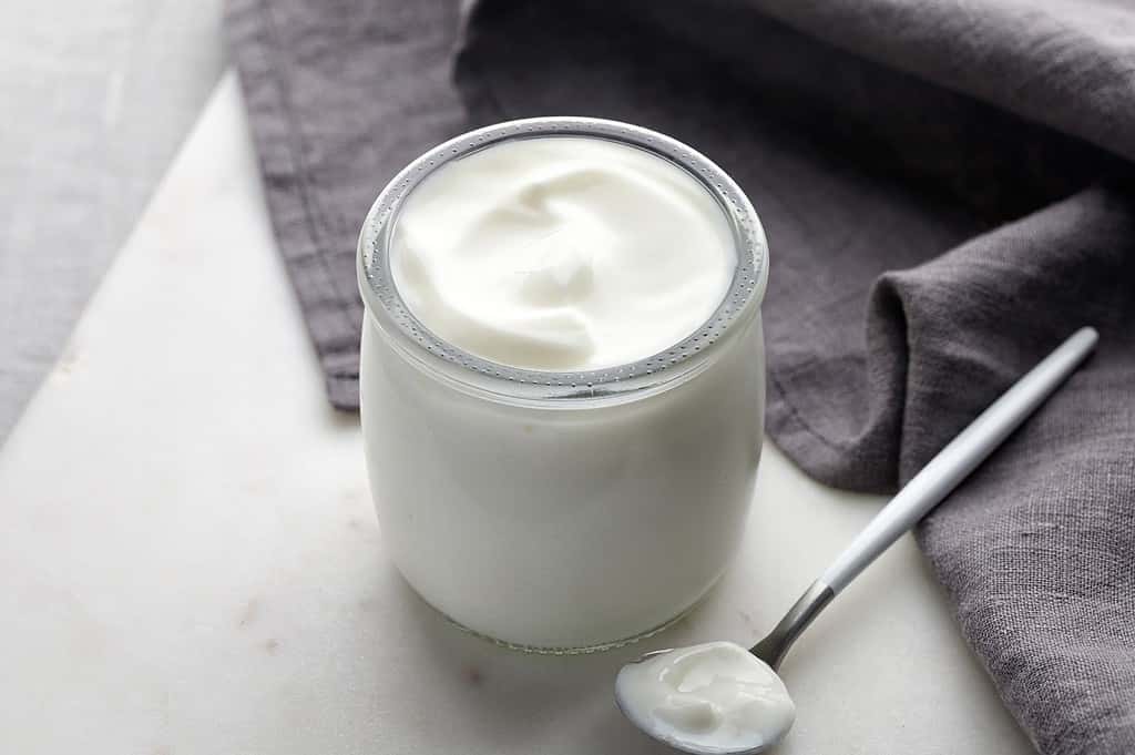A jar of fresh Greek yogurt with a spoon on a white surface, showcasing a nutritious and delicious dairy snack.
