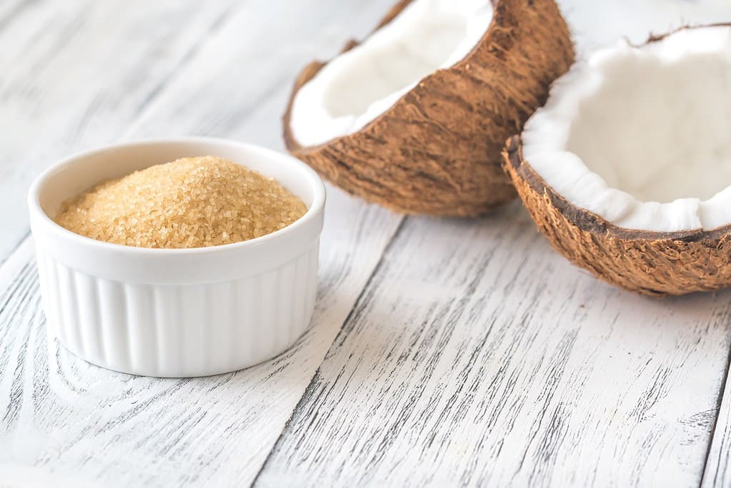 Natural coconut sugar in a white ramekin with split coconut shells on rustic wooden surface.