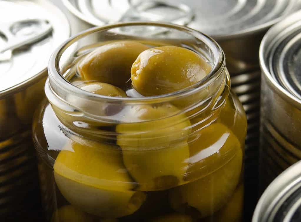 Delicious green olives in a glass jar, perfect for snacking or cooking, on a background of unopened canned goods.