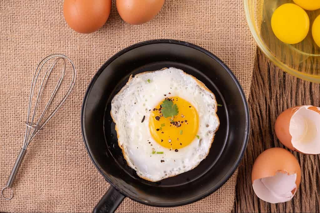 Freshly cooked sunny side up egg on a black plate, with eggshells and raw eggs nearby, ready for breakfast.