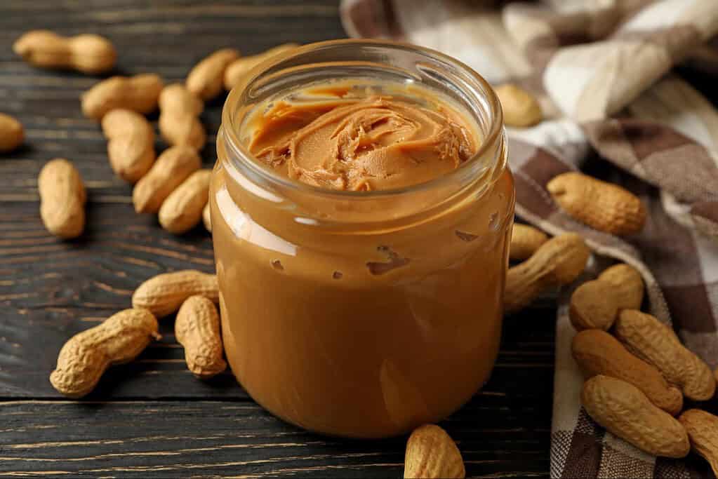 Natural peanut butter spread in a glass jar with scattered peanuts on a rustic wooden surface.