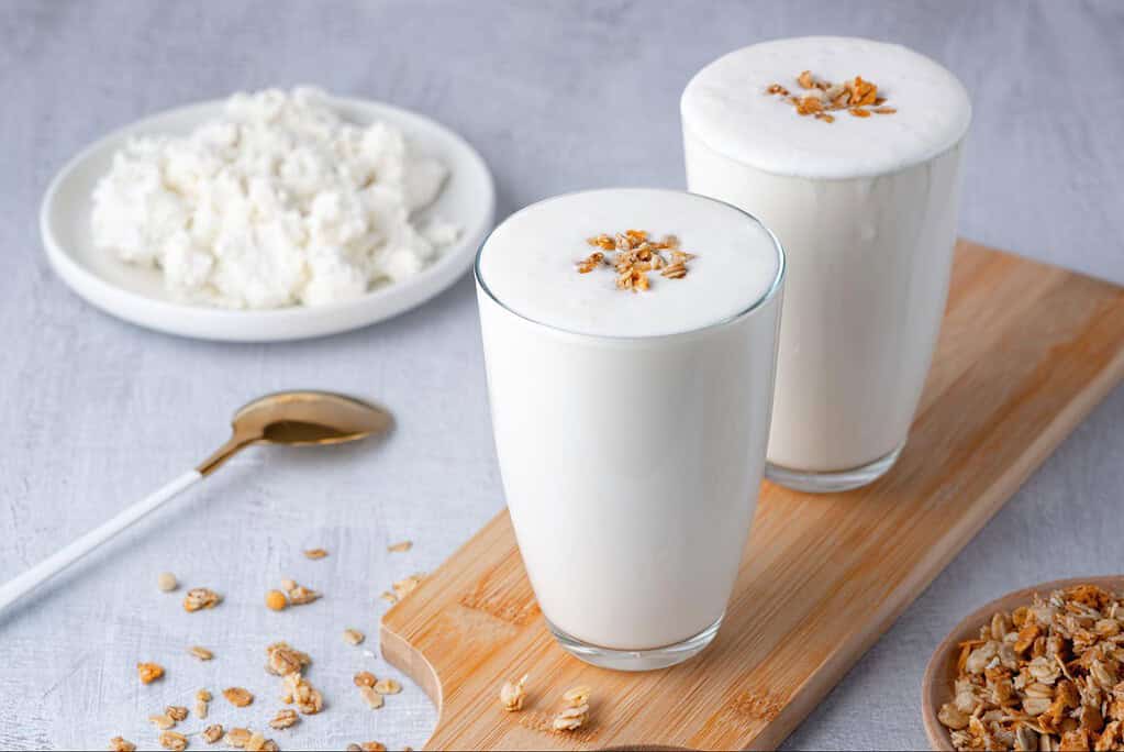 Bright image of two glasses of milk topped with oats, with oats scattered on a white surface and a plate of oats in the background.