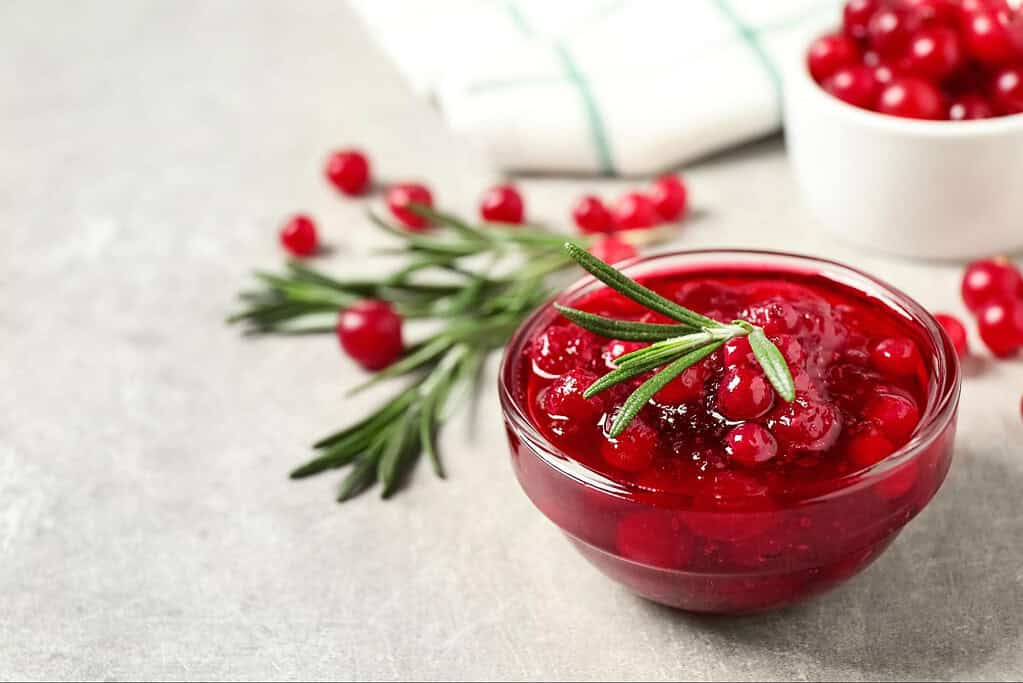 Vibrant bowl of cranberry sauce garnished with rosemary, complemented by herb sprigs and a bowl of fresh cranberries.