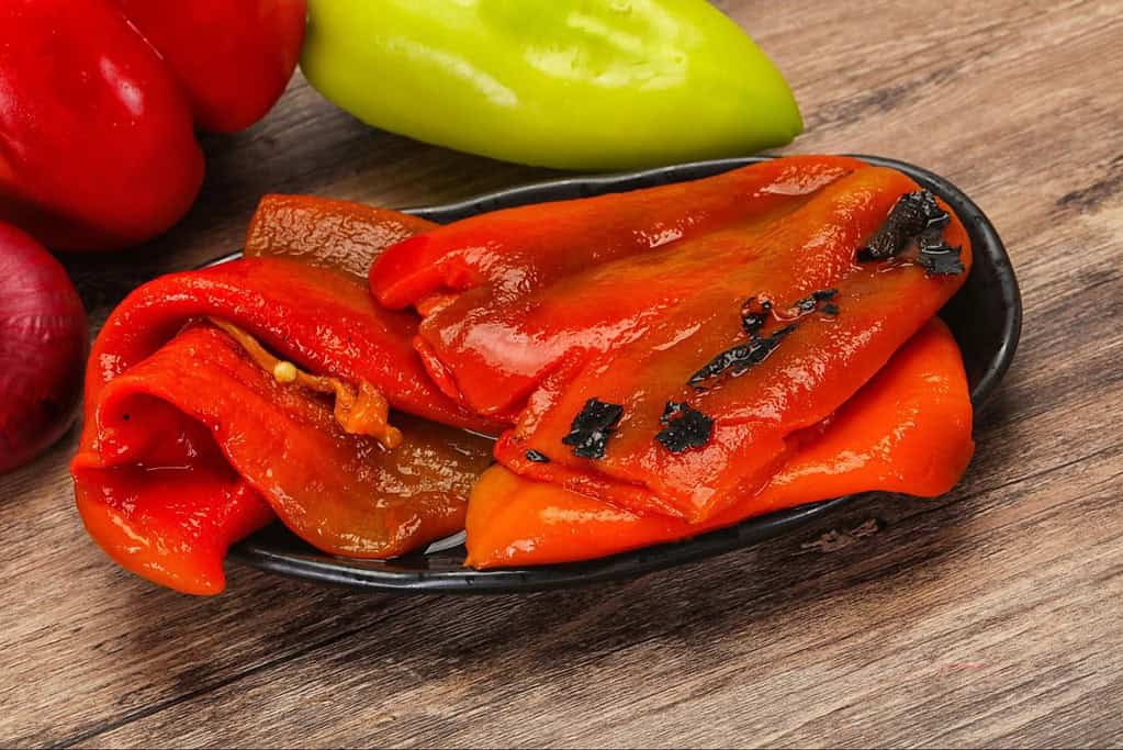 Close-up of charred red bell peppers on a black dish with fresh peppers in the background.