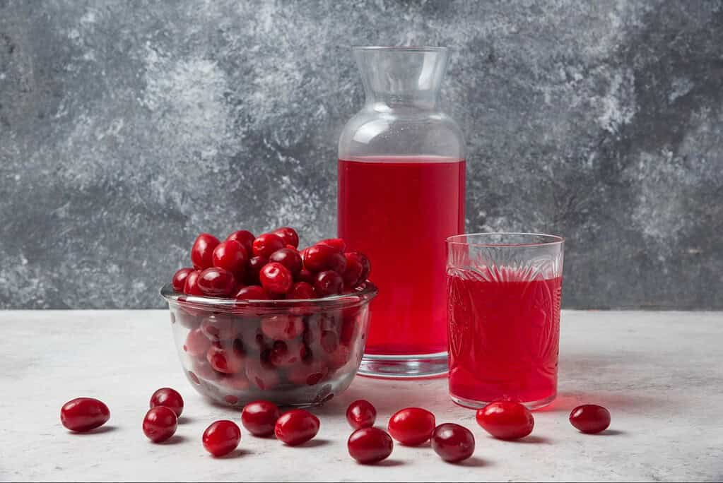 Rich cherry juice in a glass pitcher and a glass of cherry juice, alongside fresh cherries, on a white surface with a textured grey background.