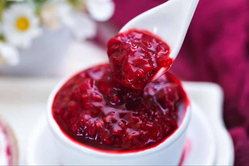 Close-up of vibrant red berry jam in a white spoon, ready for spreading or topping.