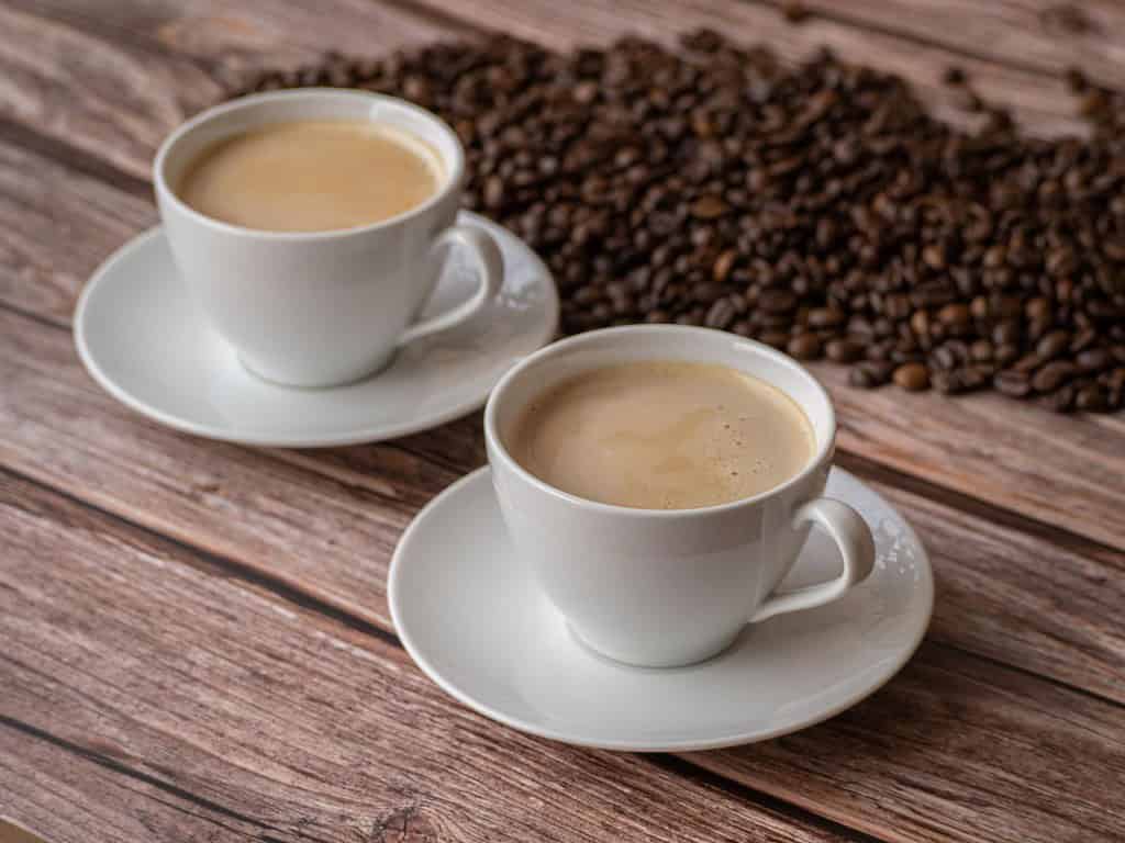 An inviting scene of two white coffee cups filled with fresh coffee, surrounded by roasted coffee beans on a rustic wooden table.