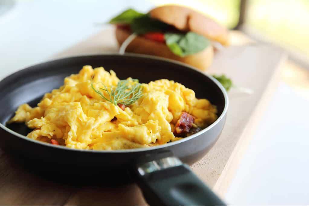 Scrambled eggs served in a black skillet, garnished with fresh herbs, with a breakfast sandwich in the background.