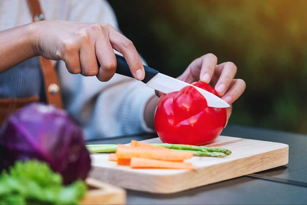 Close-up of a person slicing a red bell pepper on a wooden cutting board outdoors. Fresh vegetables ready for cooking or pet food.