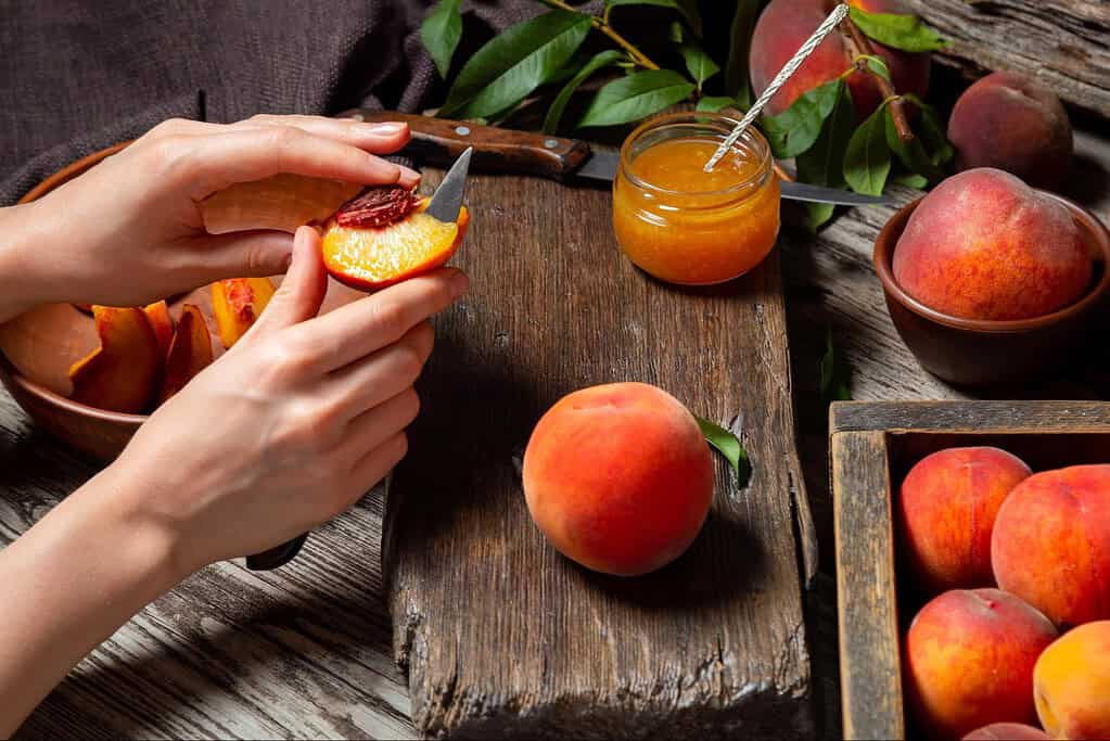 Close-up of hands preparing peach jam with fresh peaches and jam jar on rustic wooden table, healthy homemade fruit preserves.