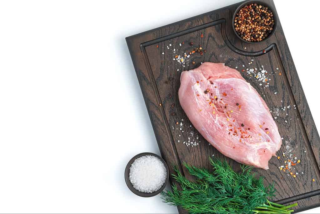 Fresh raw chicken breast seasoned with salt and pepper on a wooden cutting board with herbs and spices.