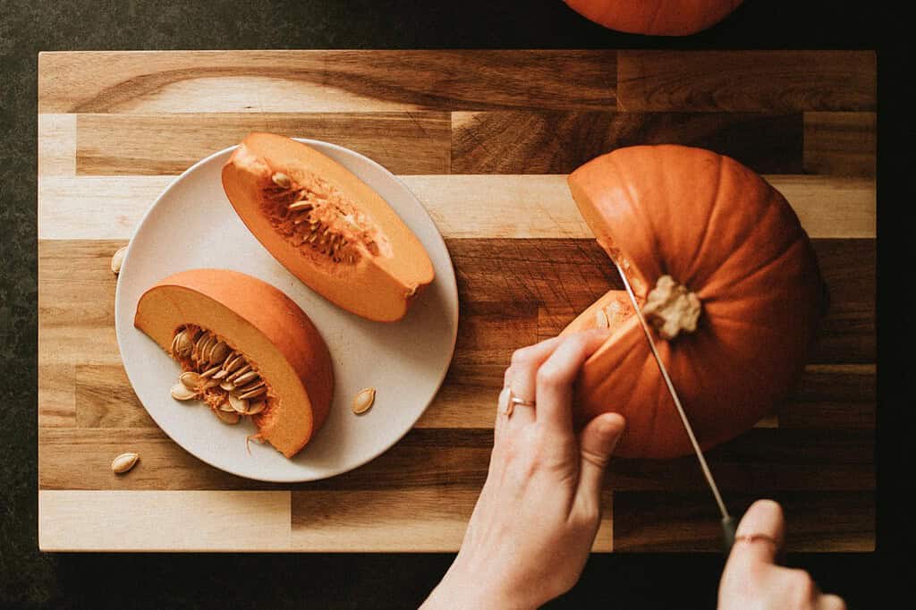 Hands slicing fresh pumpkin on wooden cutting board with pumpkin seeds.