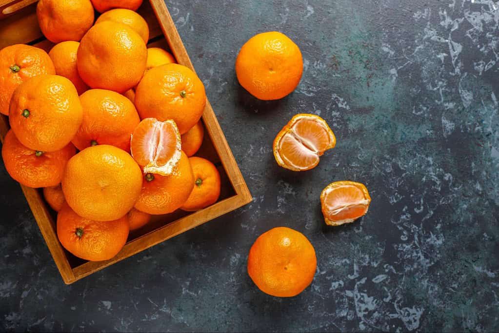 Vivid orange tangerines, some peeled, scattered on textured dark surface, highlighting fresh citrus fruits.