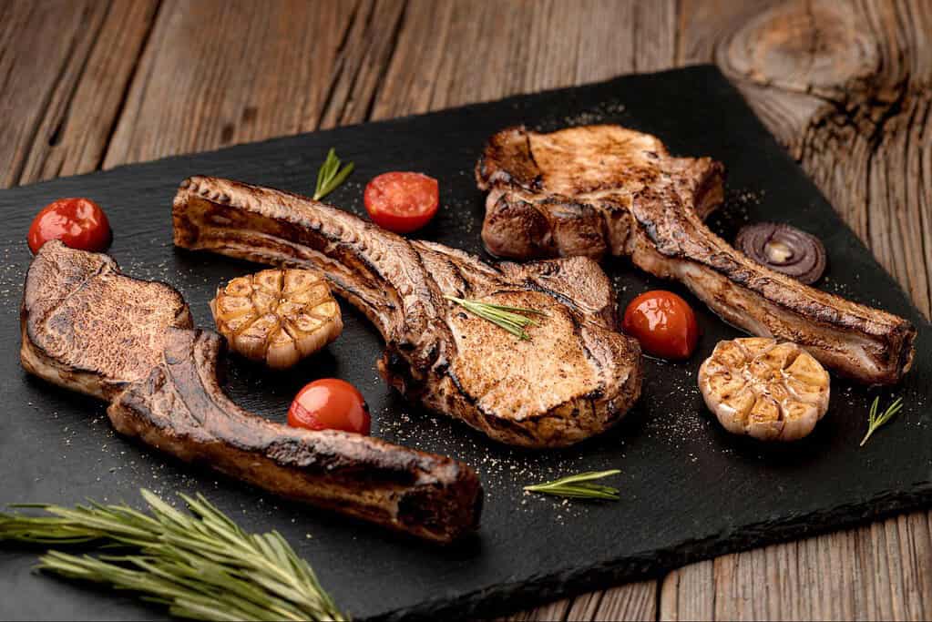 Close-up of grilled beef steaks with garlic, tomatoes, and herbs on a rustic wooden table.