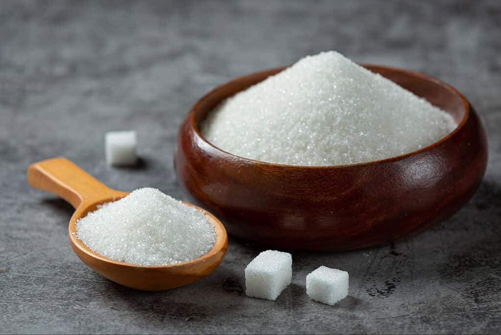 Alt text: Wooden bowl filled with white granulated sugar with sugar cubes and scoop nearby.