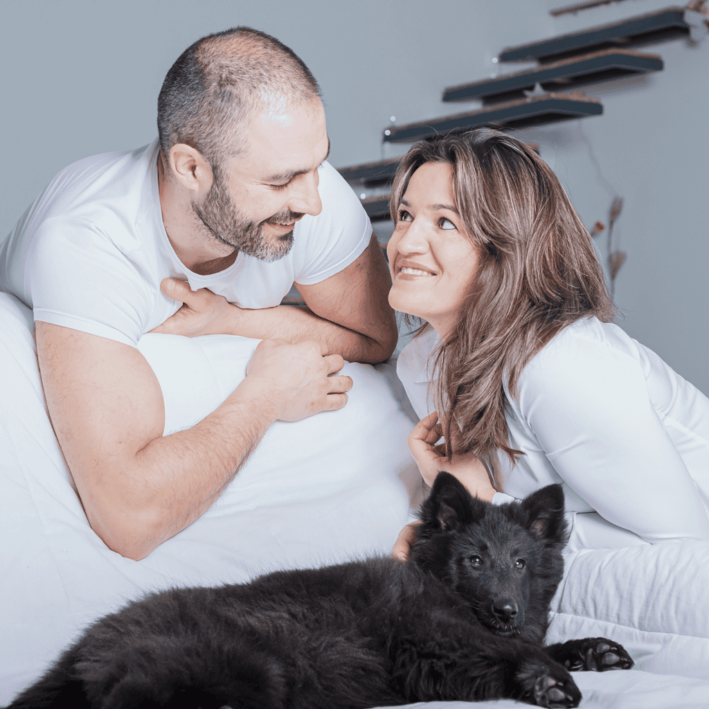 Close-up of a joyful couple with their black puppy lying on a bed, showcasing pet care and companionship.