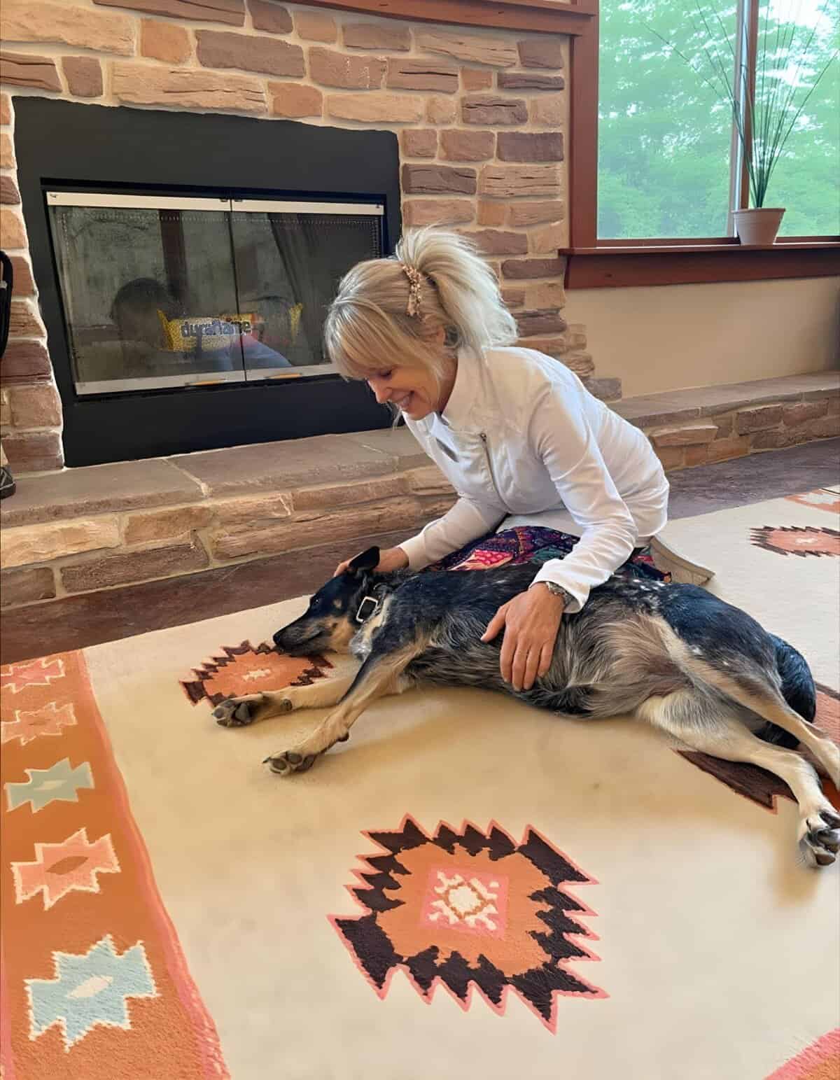 A woman happily kneels beside her dog on a colorful rug in front of a cozy fireplace indoors.