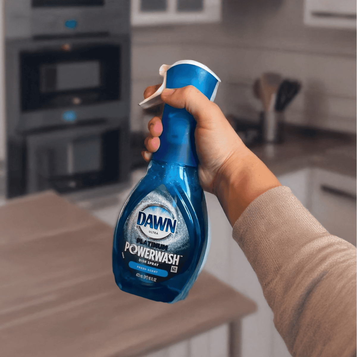 Alt text: Person holding blue Dawn Powerwash dish spray bottle in a kitchen for cleaning.