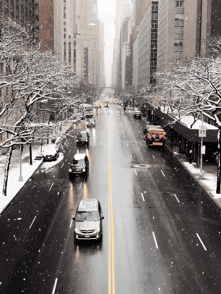 Urban winter scene with snow, tall buildings, and cars on a city street.