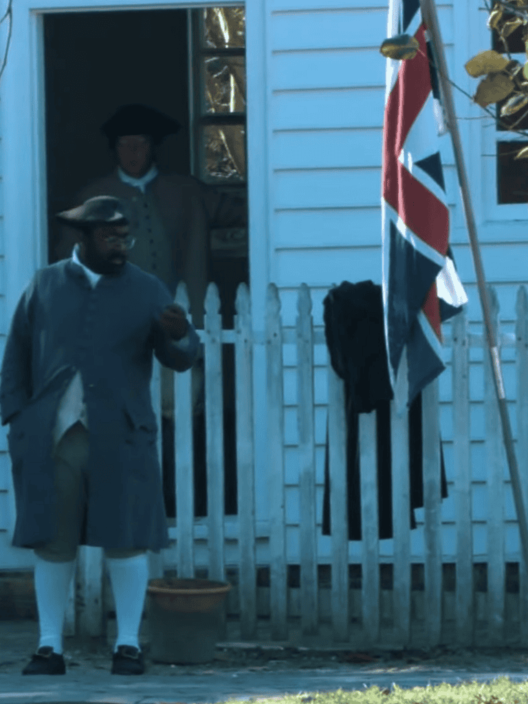 Costumed individual dressed as a colonial-era figure standing outside a house with a British flag.