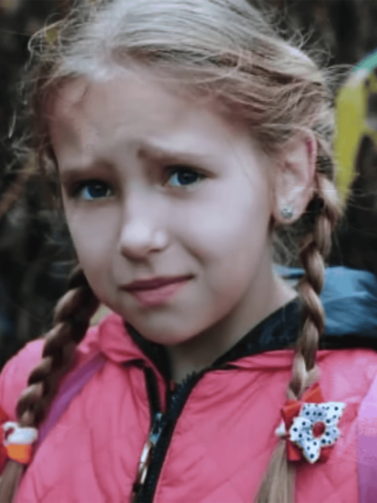 Young girl with braided blonde hair, Blue eyes, pink jacket, outdoor setting, smiling.