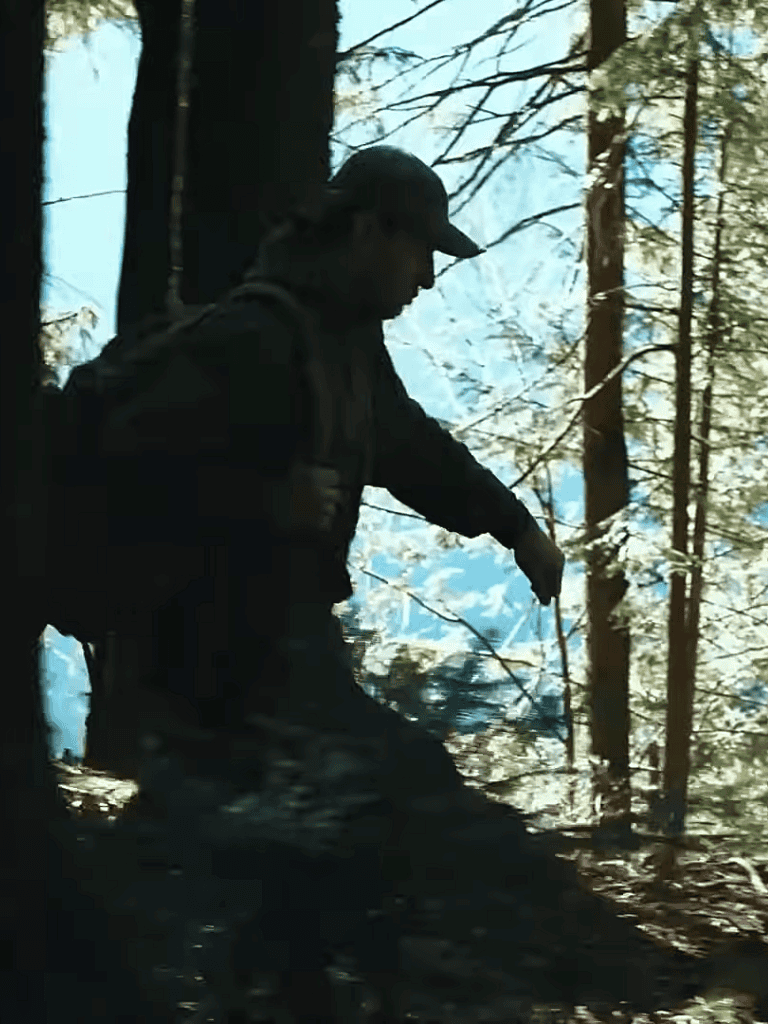 Hiker in woods with backpack and cap near trees.