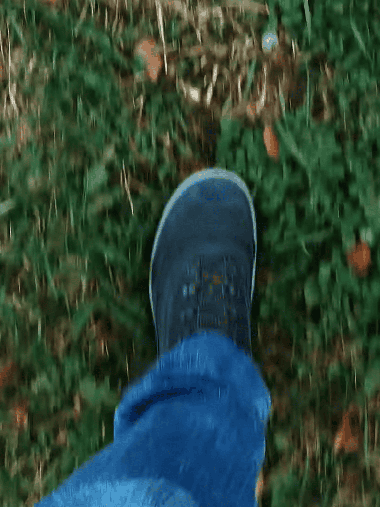 Close-up of a person's foot in a blue sneaker walking on grass and fallen leaves.