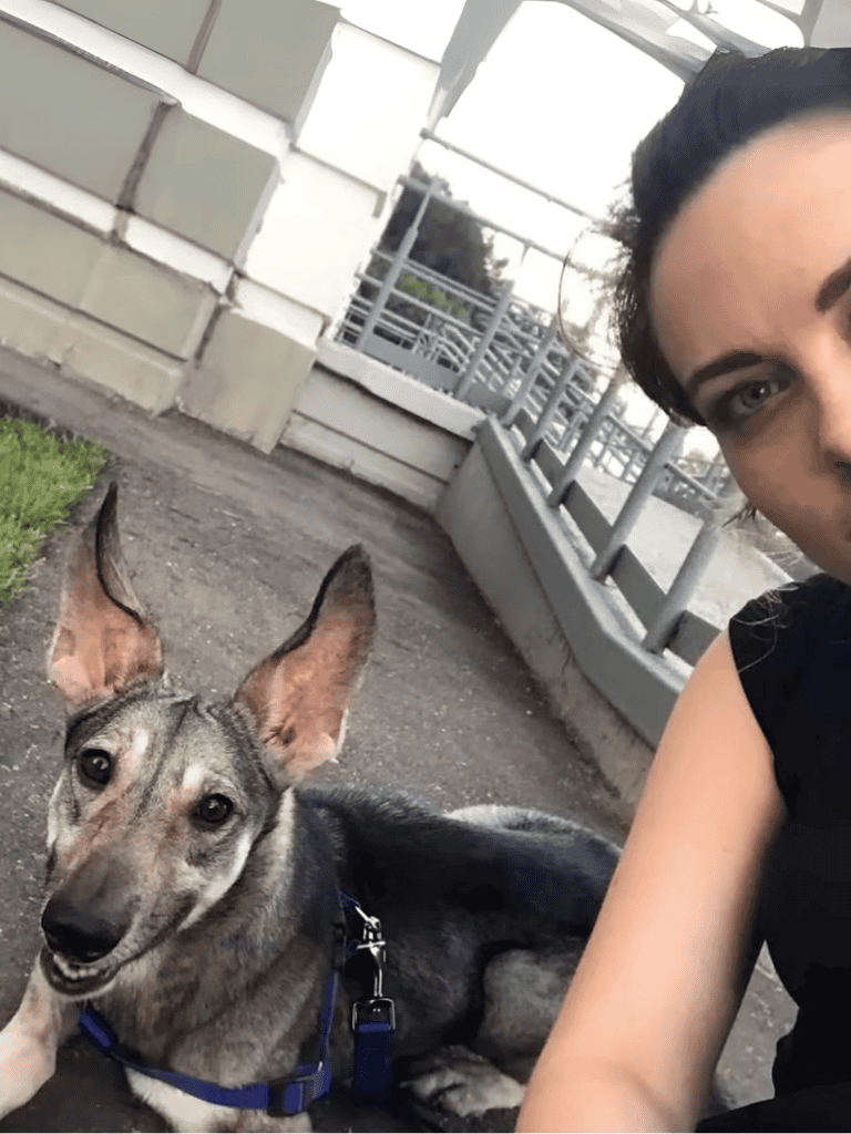 Alt text: Smiling dog lying on the ground with a woman taking a selfie outdoors.