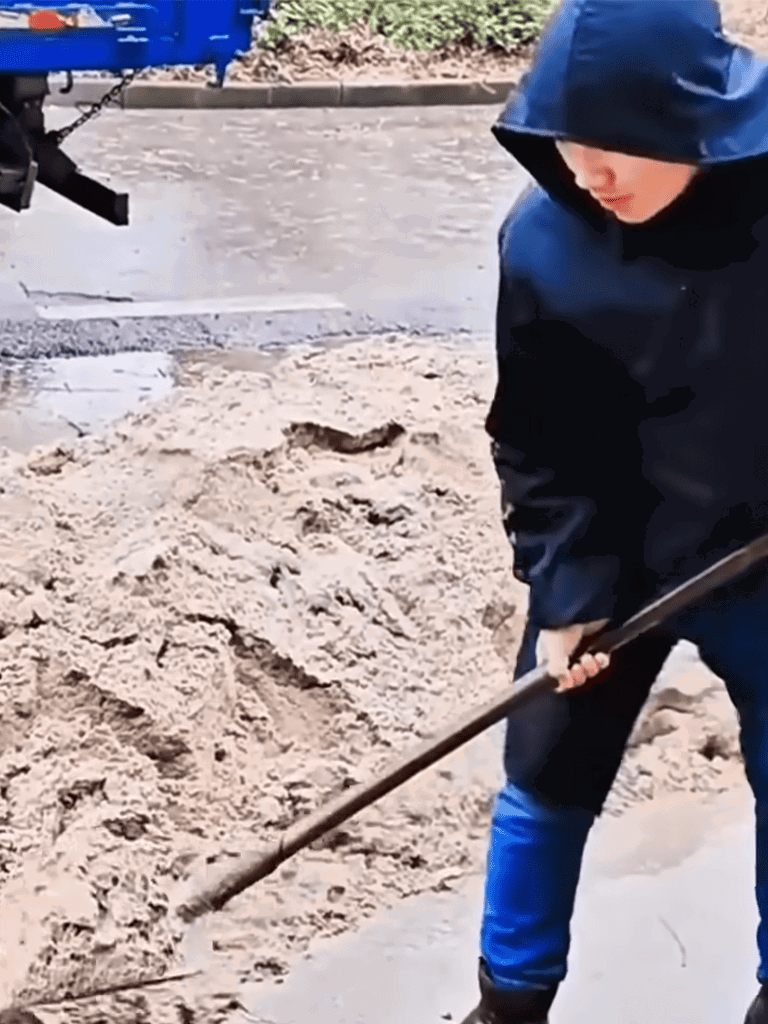 Person shoveling sand with a long-handled shovel in rainy conditions.