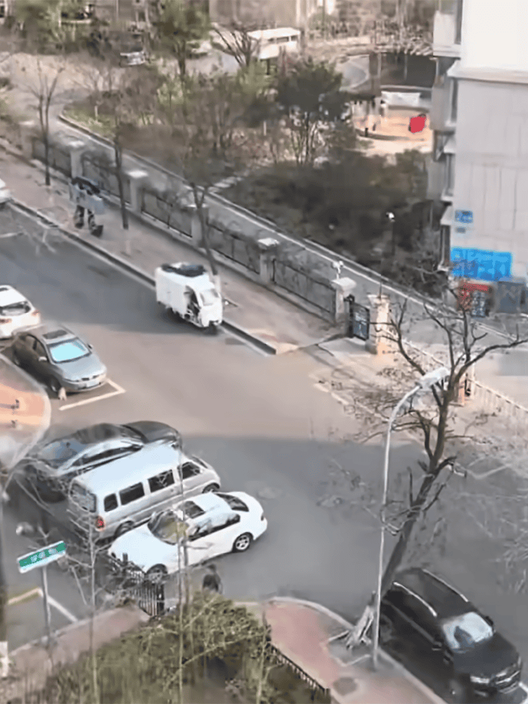 A busy urban street with parked cars, trees, and buildings, captured from above, showing city activity and infrastructure.
